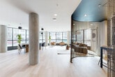 Located at 4845 Ch De La Côte-Saint-Luc in Montréal, this thoughtfully designed co-working space offers a sophisticated and functional environment for professionals. The single-floor layout is organized as an open-plan living area that seamlessly integrates distinct collaborative zones, all unified by light hardwood flooring and crisp white ceilings with recessed lighting.

A defining architectural feature is the curved room divider draped in a metallic chain-link curtain beneath a powder-blue ceiling accent, creating a semi-private lounge nook that adds visual intrigue and a sense of intimacy within the open floor plate. Exposed concrete columns punctuate the space, lending an industrial contrast to the otherwise refined, contemporary aesthetic.

Floor-to-ceiling windows wrap the perimeter, flooding the interior with abundant natural light and providing open views of the surrounding Montréal streetscape. A dedicated meeting and dining area features an oval wood table surrounded by sleek charcoal chairs, anchored by a dramatic multi-pendant black light fixture overhead. Adjacent to this zone, a fully equipped kitchenette with dark cabinetry provides practical amenities for occupants.

The lounge area is furnished with a deep sofa, leather accent chairs, a sculptural coffee table, and curated greenery, creating a welcoming atmosphere that balances productivity with comfort. The overall ambiance is modern, bright, and professionally curated.