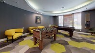 This well-appointed game room offers a dynamic and inviting space designed for social interaction and recreation. The room is anchored by a striking tray ceiling with recessed lighting and a curved crown detail that adds an elegant architectural touch to the otherwise playfully designed interior. The walls are finished in a bold charcoal gray, creating a sleek backdrop that complements the room's energetic atmosphere. A standout piece of wall art — a pineapple painting — adds a pop of personality and color to the space.

The flooring features a bold hexagonal patterned carpet in shades of yellow, gray, and taupe, tying together the room's contemporary color palette. A full-size foosball table and a billiards table serve as the centerpieces of the room, offering multiple entertainment options. Bright yellow lounge chairs and an ottoman are arranged to provide comfortable seating between games. Natural light filters in through window blinds along one wall, adding warmth to the space.

A dedicated media and storage area along one wall showcases rich, dark wood cabinetry with a long countertop and a large wall-mounted flat-screen television, ideal for gaming or streaming. The room is accessible through a standard doorway that connects to an adjacent corridor, making it a well-integrated communal amenity.