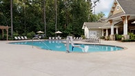 Located at 3500 Pinckney Marsh Ln in Summerville, SC, this single-story covered outdoor amenity space is designed for both relaxation and entertainment. The structure features a striking coffered ceiling with ceiling fans and integrated lighting, supported by elegant timber-frame columns that frame wide-open views of the adjacent swimming pool and surrounding wooded landscape.

The space is divided into distinct functional zones, all unified by warm terracotta tile flooring throughout. A generously sized lounge area is furnished with wicker seating and plush cushions, creating a comfortable gathering spot. Adjacent to it, a dedicated dining area accommodates a round glass-top table with seating for six, positioned near a wall-mounted flat-screen television and a built-in wet bar with dark cabinetry and sink.

A game area adds a recreational element to the space, featuring a full-size shuffleboard table and a ping pong table, making this an ideal setting for social events. A row of white-framed French doors lines the interior wall, allowing seamless indoor-outdoor flow and filling the space with natural light.

Outside, the expansive pool deck offers lounge chairs and umbrella seating, set against a backdrop of tall pines that provide natural privacy. The overall design blends Southern charm with modern outdoor living functionality.
