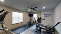 This single-floor fitness space is thoughtfully designed to provide a comprehensive workout environment. The room is finished with light-toned walls and smooth flooring, with protective rubber mats laid beneath the equipment to define the workout zone and safeguard the floor surface. Natural light fills the space through multiple windows fitted with horizontal blinds, creating a bright and energizing atmosphere. A ceiling fan with an integrated light fixture ensures steady air circulation, keeping the environment comfortable during exercise sessions. Recessed lighting supplements the natural light for consistent illumination throughout. The room is equipped with a notable selection of fitness equipment, including a treadmill, a stationary bike, and a weight bench paired with a dumbbell rack, offering options for both cardiovascular and strength training workouts. A wall-mounted flat-screen television is positioned for easy viewing during exercise, adding an entertainment element to the space. A framed piece of artwork on the wall contributes a subtle decorative touch. Overall, this fitness room balances functionality and comfort, making it a practical and inviting space for daily physical activity.