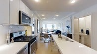 Located at 1133 Warburton Ave in Yonkers, NY, this 2-bedroom, 2.5-bathroom residence offers a thoughtfully designed single-floor layout that balances modern aesthetics with everyday functionality. The open-concept living area serves as the heart of the home, seamlessly connecting the kitchen, dining, and living spaces in one fluid arrangement. The kitchen is outfitted with stainless steel appliances — including a gas range and built-in microwave — complemented by crisp white cabinetry, subway tile backsplash, and an expansive white quartz countertop that doubles as a casual dining or prep surface. Light hardwood flooring runs throughout, lending warmth and continuity across the space. The dining area features a round table with stylish gold-accented chairs, flowing naturally into the living room, which is anchored by a blue sofa and bathed in natural light from large windows at the far end. The windows frame what appears to be an open, bright exterior view, adding a sense of openness and airiness to the interior. Recessed lighting throughout ensures a well-lit environment at any time of day. The overall ambiance is clean, contemporary, and inviting — ideal for both comfortable daily living and entertaining.
