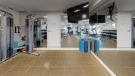 This fitness center is a thoughtfully designed, single-level space offering a comprehensive workout environment. The room is elongated and open, with durable tile flooring throughout and recessed fluorescent lighting that ensures consistent illumination across the entire area. Large windows line one wall, flooding the space with natural light and providing views of the surrounding wooded and landscaped scenery, creating an energizing atmosphere for exercise.

The cardio zone features multiple treadmills positioned to face the windows, allowing users to enjoy outdoor views during workouts. Several elliptical cross-trainers occupy additional floor space, complementing the cardio offerings. A wall-mounted flat-screen television is centrally positioned for entertainment during exercise sessions. A mini-split climate control unit is installed on the wall, ensuring comfortable temperatures year-round.

The strength training area includes a well-stocked dumbbell rack with a full range of weights, a multi-function cable machine, and a flat weight bench. Branded yoga mats are neatly stored near the mirrored wall, which spans a significant portion of the room, visually expanding the space and supporting form-checking during workouts. Motivational phrases such as "Wake. Run." and "Sleep. Repeat." are displayed as bold wall decals, reinforcing the space's active, goal-oriented ambiance.
