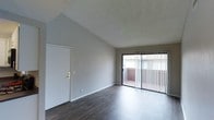 This single-story space showcases a clean, open layout highlighted by striking vaulted ceilings that add a sense of height and airiness to the interior. The living area features dark hardwood-style flooring that contrasts elegantly with the neutral gray walls, creating a modern yet inviting atmosphere. A textured ceiling adds subtle visual interest overhead.

Natural light flows generously into the space through a large sliding glass door fitted with vertical blinds, which opens to an outdoor balcony or patio area — a functional extension ideal for relaxing or entertaining. The sliding door provides pleasant outdoor views and enhances the sense of openness throughout the main living area.

The entry door is positioned along the adjacent wall, offering a practical and straightforward layout. Clean white trim and baseboards frame the space neatly, complementing the overall minimalist aesthetic. The combination of vaulted ceilings, natural light from the sliding glass access point, and the warm tones of the flooring give this space a welcoming and well-balanced ambiance suitable for a variety of furnishing styles.