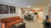 This single-floor residence offers a comfortable and practical layout built around an open-concept living space. The living area is anchored by plush wall-to-wall carpeting in a warm neutral tone, providing a soft and inviting atmosphere. A cozy sofa in a rich terracotta hue serves as the focal point of the lounge area, complemented by a wicker coffee table and a classic floor lamp that adds a warm ambient glow.

The living area flows seamlessly into the dining space, where a round glass-top table surrounded by dark wood chairs with upholstered seats creates an elegant yet casual setting. Framed artwork adorns the walls, adding personality and a finished look to the shared living and dining zone.

Just beyond the dining area, a half-wall peninsula separates the kitchen, which features warm wood-toned cabinetry, a built-in overhead light fixture, and essential appliances including a refrigerator. The kitchen layout is efficient and well-organized, offering ample counter and storage space.

Throughout the space, the consistent use of neutral wall tones, soft carpeting, and warm lighting creates a cohesive, welcoming ambiance. The open flow between the living, dining, and kitchen areas makes the space feel connected and versatile, ideal for both everyday living and casual entertaining.