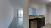 This single-floor space offers a clean, modern aesthetic defined by an open-concept layout and an abundance of natural light. The living area features smooth hardwood-style flooring and expansive floor-to-ceiling windows dressed with vertical blinds, allowing daylight to fill the space while framing outdoor views. The bright, airy atmosphere is enhanced by crisp white walls and a simple, unfussy design that maximizes the sense of openness.

The kitchen is positioned adjacent to the living area and is partially separated by a structural column, creating a natural division while maintaining visual connectivity throughout the unit. It is equipped with granite countertops featuring speckled detailing, a stainless steel sink, and stainless steel appliances, including a dishwasher. Dark cabinetry provides a contrasting accent against the light tones of the surrounding space.

Overall, the unit presents a contemporary and functional layout well-suited for everyday living. The interplay of natural light, neutral finishes, and modern fixtures creates a welcoming and practical environment. The open sightlines between the kitchen and living area contribute to a cohesive, spacious feel throughout the entire floor.