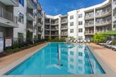 This amenity space is designed to offer residents a comprehensive recreational experience across two distinct areas. The outdoor pool area is the centerpiece of the complex, framed by a multi-story residential building with private balconies. The generously sized lap pool features clean blue water, a surrounding pool deck furnished with lounge chairs and shaded seating areas, and lush landscaping that enhances the resort-like atmosphere.

Inside, the amenity lounge is divided into a game room and a social lounge. The game room is visually striking, featuring a bold textured dark-tile accent wall that serves as a dramatic backdrop for a full-size billiards table and a shuffleboard table. Geometric gold pendant lights hang overhead, adding a modern, upscale touch. A dining table provides additional space for gatherings.

The adjoining lounge area is furnished with plush, upholstered armchairs arranged around a wall-mounted flat-screen television, supported by built-in dark wood cabinetry. A patterned carpet in deep, rich tones grounds the space and adds warmth. Floor-to-ceiling drapes frame glass doors leading to the outdoor area, allowing natural light to filter in and connecting the interior to the pool beyond.
