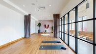 Located at 440 W State St in Columbus, Ohio, this yoga and meditation studio offers a thoughtfully designed space for wellness and mindful practice. The single-floor layout provides an open, unobstructed practice area finished with warm-toned hardwood flooring that runs throughout, contributing to a grounded and inviting atmosphere.

A defining architectural feature is the expansive floor-to-ceiling steel-framed glass wall along one side of the studio, flooding the interior with abundant natural light and offering views of the surrounding urban environment. Tall, flowing taupe curtains are mounted on ceiling tracks and can be drawn to divide the space or create a more intimate, private setting.

The focal point of the meditation area is a mauve-toned accent wall adorned with a graceful textile wall hanging, flanked by lush potted plants and decorative crystal arrangements that enhance the tranquil ambiance. Exposed concrete columns add an industrial character that contrasts beautifully with the soft, neutral color palette.

Additional features include recessed ceiling lighting, a built-in storage cabinet with open shelving for equipment, and a cushioned floor seating arrangement. The studio is equipped with yoga mats and is well-suited for group classes, private sessions, or guided meditation. The overall design balances modern minimalism with warm, organic elements to create a calming and functional wellness environment.