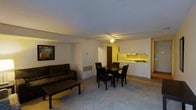 This single-floor space is designed around an open-concept layout that seamlessly connects the living, dining, and kitchen areas into one cohesive environment. The living area is carpeted and comfortably accommodates a dark upholstered sofa paired with a rectangular coffee table, creating a relaxed seating space. A framed piece of wall art adds a decorative touch to the neutral-toned walls. Overhead, a ceiling fan with a light fixture provides both ventilation and ambient lighting throughout the main living space. A wall-mounted heating unit is also visible, contributing to the room's practical amenities. Adjacent to the living area, the dining space features a dark wood rectangular table surrounded by four matching chairs, offering ample room for meals and gatherings. The kitchen is partially open to the main living area via a pass-through counter, and is finished with light-colored cabinetry and under-cabinet lighting that gives the space a warm glow. A door visible in the background likely leads to a secondary room or utility area. The overall ambiance is warm and functional, with neutral tones and carpeted flooring throughout.