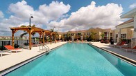 This outdoor amenity area centers around a generously sized swimming pool with clear, blue-tinted water, designed to evoke a resort-like atmosphere. The pool deck is finished in light-colored concrete, providing ample space for relaxation and movement around the water. A prominent wooden pergola anchors one side of the deck, offering a shaded retreat and serving as a distinctive architectural focal point with its warm, natural timber construction. Surrounding the pool, rows of lounge chairs with red cushions invite residents to enjoy the open-air setting, while red-accented umbrella tables provide additional shaded seating nearby. Decorative pole-mounted lanterns are positioned throughout the deck, enhancing the ambiance during evening hours. Lush palm trees are planted throughout the area, reinforcing a tropical aesthetic and providing natural shade. The backdrop consists of two-story residential buildings with warm yellow and tan exteriors and pitched rooflines, creating a cohesive and inviting community setting. The space is open to the sky, allowing for abundant natural light throughout the day, and the overall design prioritizes outdoor living, leisure, and a vibrant community atmosphere.