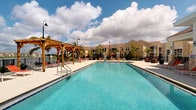 This outdoor amenity area centers around a generously sized swimming pool with clear, blue-tinted water, designed to evoke a resort-like atmosphere. The pool deck is finished in light-colored concrete, providing ample space for relaxation and movement around the water. A prominent wooden pergola anchors one side of the deck, offering a shaded retreat and serving as a distinctive architectural focal point with its warm, natural timber construction. Surrounding the pool, rows of lounge chairs with red cushions invite residents to enjoy the open-air setting, while red-accented umbrella tables provide additional shaded seating nearby. Decorative pole-mounted lanterns are positioned throughout the deck, enhancing the ambiance during evening hours. Lush palm trees are planted throughout the area, reinforcing a tropical aesthetic and providing natural shade. The backdrop consists of two-story residential buildings with warm yellow and tan exteriors and pitched rooflines, creating a cohesive and inviting community setting. The space is open to the sky, allowing for abundant natural light throughout the day, and the overall design prioritizes outdoor living, leisure, and a vibrant community atmosphere.