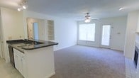 This single-floor residence offers a clean, open-concept layout that seamlessly connects the kitchen and living areas. The kitchen features a raised bar counter with dark granite-style countertops and an integrated sink, providing both functional workspace and a casual dining or entertaining surface. Adjacent built-in shelving adds convenient storage and display space, enhancing the room's utility without sacrificing style.

The living area is finished with wall-to-wall carpeting in a neutral tone, creating a warm and comfortable atmosphere throughout. A ceiling fan with a light fixture is centrally positioned overhead, ensuring year-round comfort and ambient lighting. Large windows along the exterior wall flood the space with generous natural light, contributing to the bright and airy feel of the interior. A door provides direct access to the outdoors, further enhancing the connection between interior and exterior living.

The walls are painted in a crisp, neutral white, offering a versatile backdrop that complements a wide range of furnishing styles. Overall, this space is characterized by its practical design, thoughtful built-in features, and an inviting, light-filled ambiance well-suited for everyday living.