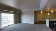 This contemporary apartment showcases a well-designed, open-concept living space. The room is characterized by its generous dimensions and thoughtful layout. A standout feature is the large sliding glass door, which not only allows ample natural light to flood the space but also potentially offers access to an outdoor area, enhancing the apartment's livability.

The interior boasts a neutral color scheme, with soft grey carpeting covering the entire floor, providing both comfort and style. Clean white walls contribute to the bright, airy atmosphere, while a subtle grey accent wall adds depth and visual interest.

The ceiling design is notable, with a slight elevation in one section, creating a sense of increased height and spaciousness. This architectural detail adds character to the otherwise minimalist space.

An entryway leads into the apartment, marked by a yellow door visible in the distance, creating a clear delineation between public and private spaces. Adjacent to this area, there appears to be additional rooms or perhaps a kitchen, hinting at a well-planned, functional layout.

Lighting is strategically placed, with a ceiling fixture in the entryway providing warm illumination. The room's design allows for flexible furniture arrangement, making it adaptable to various lifestyle needs. Overall, this apartment presents a blank canvas, ready for personalization while offering modern comforts and aesthetic appeal.