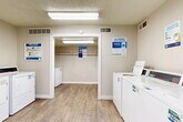 This shared laundry facility is a practical, well-organized space designed to accommodate multiple users efficiently. The room features durable wood-look flooring throughout, providing a clean and easy-to-maintain surface that lends a modern touch to the functional environment. Bright fluorescent ceiling fixtures ensure the space is well-lit at all times. The layout is thoughtfully arranged, with multiple commercial-grade top-load washing machines and dryers positioned along the walls, offering ample capacity for residents. A dedicated alcove area provides additional machine placement and features a wall-mounted hanging rod, ideal for garments that require air drying. Instructional signage and usage guides are posted throughout, making the facility user-friendly. A prominent French door entrance with glass panes allows natural light to filter in and provides a view of the surrounding brick exterior and greenery. A fire extinguisher and safety signage near the entrance reflect attention to occupant safety. A community bulletin board and informational notices contribute to the welcoming, organized atmosphere. Ventilation is supported by ceiling-mounted HVAC vents, keeping the space comfortable during use. The neutral beige walls and textured ceiling tie the room together, creating a clean and functional laundry environment.