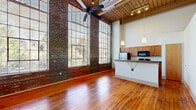 Located at 2100 Main Street in Porterdale, GA, this one-bedroom, one-bathroom loft-style unit showcases the character of an adaptively reused industrial building. The open living area is the centerpiece of the space, boasting soaring ceilings with exposed wooden beams, original hardwood flooring, and dramatic floor-to-ceiling multi-pane windows that flood the interior with natural light and frame views of the surrounding greenery. A ceiling fan and track lighting complement the industrial aesthetic, while exposed ductwork adds to the loft's authentic charm. The kitchen features warm wood cabinetry, granite-style countertops, a breakfast bar peninsula, and full appliances including a stainless steel refrigerator and gas range. A hallway with polished concrete flooring connects the main living area to the private bedroom, which includes a narrow vertical window and soaring ceilings. A dedicated closet provides convenient storage. The full bathroom is finished with tile flooring, a tiled tub-shower surround, a wood vanity, and a round mirror. Throughout the unit, a thoughtful contrast between polished concrete, rich hardwood, and exposed brick creates a cohesive, characterful industrial-chic ambiance.