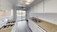 This single-floor space offers a clean, functional layout with a cohesive design aesthetic carried throughout. The living area is bright and open, featuring wood-style flooring in a cool gray tone that extends seamlessly across the unit. Natural light streams in through large windows dressed with vertical blinds, casting a warm glow across the floors. A wall-mounted HVAC unit ensures year-round comfort in the main living space.

The layout connects the living area to the kitchen and additional rooms through open passageways, with a subtle stepped platform detail adding architectural interest to the transition zones. A tall vertical heating unit is centrally positioned between the openings, serving as a functional design element.

The kitchen is well-appointed with white cabinetry, granite-style countertops, a double stainless steel sink, and a gas range. A ceiling fan provides additional ventilation and airflow. A sliding glass door at the end of the kitchen opens to an outdoor area, filling the space with natural light and offering convenient exterior access.

The bedrooms are generously sized with textured ceilings, vertical blind-covered windows that bring in ample daylight, and the same wood-style flooring found throughout the unit. The overall ambiance is light, neutral, and move-in ready, with a practical layout suited for comfortable everyday living.