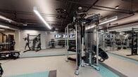 Located at 425 Hillsborough St in Chapel Hill, NC, this comprehensive fitness center is designed to support a full range of workout disciplines within a single, well-organized floor. The space features an industrial-modern aesthetic, with exposed ductwork and dark ceilings contrasted by bright linear LED lighting and teal accent walls that lend the facility a dynamic, energetic atmosphere.

The cardio zone is lined with a row of treadmills and elliptical machines, complemented by rowing machines and additional aerobic equipment, all positioned across color-blocked rubber flooring in neutral and teal tones. A dedicated strength training area offers an extensive dumbbell rack spanning a wide weight range, multiple benches, a power rack, cable machines, and plate-loaded strength equipment — all accompanied by wall-mounted flat-screen TVs for entertainment during workouts.

A separate studio space features light wood-look flooring, pendant lighting, a full mirrored wall, and ample open floor space suited for yoga, stretching, or group fitness classes. This room is stocked with stability balls, medicine balls, resistance bands, and a central storage unit, along with an interactive fitness display screen.

Throughout the facility, motivational athletic silhouette graphics accent the walls, reinforcing the space's fitness-forward identity. The layout is thoughtfully zoned to allow simultaneous use across disciplines without crowding.