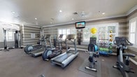This exercise room offers a dedicated fitness environment designed to support a wide range of workout routines. The space is outfitted with multiple treadmills and an elliptical trainer, providing ample options for cardiovascular exercise. A rowing machine adds versatility to the cardio offerings, while an assortment of stability balls, resistance bands, and fitness mats cater to strength and flexibility training needs. A wall-mounted flat-screen television keeps users entertained during workouts, complemented by a wall clock for convenient time tracking. The room features striped accent walls in neutral gray tones, recessed lighting, and ceiling fans that contribute to a comfortable and well-ventilated atmosphere. Large shuttered windows allow natural light to filter in while maintaining privacy, creating a bright yet focused workout environment. Informational postings and a fire safety station are also present, reflecting attention to both functionality and safety. The durable carpet flooring and protective equipment mats ensure a practical and user-friendly layout throughout the space.