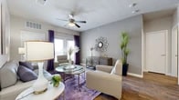 This thoughtfully designed 1-bedroom, 1-bathroom residence offers a comfortable and modern living experience across a single floor. The open-concept layout connects the kitchen, dining, and living areas seamlessly, creating an inviting flow throughout the space.

The living room is a standout feature, showcasing warm hardwood-style flooring, neutral gray walls, and a ceiling fan with light fixture. Natural light filters in through windows dressed with bold purple drape accents, adding a touch of color and personality.

The kitchen is well-equipped with rich dark cabinetry, granite-style countertops, stainless steel appliances, a tile backsplash, and a peninsula with bar seating. An adjacent dining nook with industrial-style bar-height table and stools provides a casual spot for meals.

The bedroom offers plush carpeting, a ceiling fan, and a calm, neutral palette that promotes restful living. A dedicated closet provides organized storage.

The bathroom features a dark vanity with granite-style countertop, a large mirror, wood-look flooring, and a tiled tub-shower combination — balancing practicality with a polished aesthetic.

Throughout the residence, consistent use of hardwood-style flooring, neutral tones, and modern fixtures creates a cohesive, contemporary atmosphere that is both functional and visually appealing.