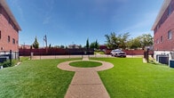 This outdoor space presents a thoughtfully designed courtyard set between several multi-story brick buildings, offering a communal green area that is both functional and visually appealing. The grounds are covered in vibrant, well-kept artificial turf that maintains a consistently lush appearance. A defining architectural feature of the courtyard is a circular concrete hardscape element at its center, connected by a straight concrete pathway that bisects the lawn and provides clear pedestrian access throughout the space.

The surrounding buildings are constructed of classic red brick, lending the area a traditional and established character. Mature trees and neatly trimmed evergreen shrubs are placed throughout the perimeter, adding natural texture and seasonal greenery. A chain-link perimeter fence borders the rear of the space, offering enclosure while maintaining sightlines to adjacent streets and neighboring structures.

An American flag is visible on a flagpole near the fencing, contributing a civic presence to the setting. The front of the property features a sloped grass lawn with concrete walkways and iron railings leading to building entrances, complemented by stone facade accents on select structures. Under open skies and abundant natural light, this courtyard serves as an inviting, open-air common area with a clean, orderly ambiance.