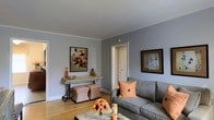 This single-floor space offers a comfortable and thoughtfully designed interior that balances style with practicality. The living area serves as the focal point, featuring smooth gray walls that create a calm, neutral backdrop, complemented by warm hardwood flooring that adds natural character throughout. Crown molding frames the ceiling, lending an elegant architectural detail to the room.

The living area is furnished with a plush gray sofa accented by vibrant orange throw pillows, creating a welcoming and warm atmosphere. A rustic console table with wrought-iron legs lines one wall, accessorized with decorative vases and framed botanical artwork that echoes the earthy color palette. Two patterned ottomans in rust and beige tones provide flexible seating and add texture to the space.

A central coffee table anchors the seating arrangement, styled with candles and a floral centerpiece featuring sunflowers that reinforce the warm, autumnal aesthetic. A doorway visible from the main living area suggests additional rooms beyond, offering functional flow throughout the floor plan.

Natural light fills the space, enhancing the warmth of the hardwood floors and the soft gray tones of the walls. Overall, this space presents a cohesive, inviting ambiance with carefully chosen décor that reflects a harmonious blend of comfort and style.