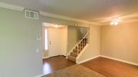 This two-story residence offers a well-organized layout with thoughtful architectural details throughout. The main level welcomes visitors through a front entryway framed by a sidelight window that allows natural light to filter in, creating a bright and inviting entry point. Crown molding runs along the upper edges of the walls, adding a refined touch to the interior, while textured ceilings lend a classic character to the space.

The main living area features rich hardwood flooring that extends through the entry and main zones, transitioning to plush carpeting in the lounge area — a combination that adds both warmth and visual interest. Neutral, earthy wall tones create a calm and versatile backdrop, suitable for a variety of décor styles.

A central staircase with white-painted balusters and wooden treads connects the main floor to the upper level, serving as both a functional and focal element of the interior. A thermostat is conveniently positioned near the entry, and a wall vent indicates a central HVAC system throughout the home.

The overall ambiance is clean, understated, and move-in ready, with consistent trim work and a cohesive neutral palette tying the spaces together for a comfortable, livable feel across both floors.