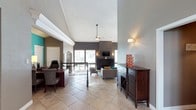This single-story space showcases an open, flowing layout enhanced by dramatically vaulted ceilings that create a strong sense of spaciousness and airiness throughout. The interior is unified by light-toned tile flooring that extends seamlessly across the entire space, contributing to a clean and cohesive aesthetic.

A dedicated work or reception area near the entrance features a rich, dark wood desk paired with a cushioned chair, offering a practical and professional zone. Adjacent to this, dark wood storage cabinetry and a wine rack credenza add both function and warmth to the space.

The main living area is anchored by a dark-painted accent wall housing a wall-mounted television and a classic fireplace, flanked by decorative wall sconces that provide warm ambient lighting. A ceiling fan with a pendant-style fixture serves the central seating arrangement, which includes a sofa, armchairs, and colorful accent pillows that inject personality into the neutral palette.

Large windows along the back wall allow natural light to filter in, while a railing detail hints at a transitional zone leading toward an outdoor area. The overall ambiance is welcoming, polished, and well-appointed.