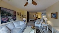 This single-floor residence offers an open, flowing layout that connects the living area, dining space, and kitchen in a cohesive and functional arrangement. The living room is furnished with a light blue upholstered sofa accented by decorative pillows, paired with a floral-patterned armchair that adds a classic, cottage-inspired charm. A white-painted coffee table serves as a central focal point, styled with decorative accessories and candles. Ceiling fans are present in both the living and dining areas, providing practical air circulation while complementing the relaxed aesthetic of the space. The walls, painted in a soft yellow tone, create a warm and welcoming ambiance throughout. Framed artwork and wall décor add visual interest and personality to the living area. The kitchen, visible from the main living space, features white cabinetry and appliances, maintaining a clean and bright appearance. A dark-toned hallway leads further into the home, suggesting additional private rooms beyond the main living zone. The flooring transitions subtly across areas, with a patterned area rug anchoring the seating arrangement. Overall, the space presents a comfortable, well-appointed interior with a traditional and homey character.