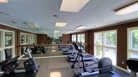 This fitness center is designed to support a well-rounded exercise routine in a comfortable, functional setting. The space is outfitted with a variety of cardio equipment, including treadmills, elliptical trainers, and stationary bikes, all arranged to allow for easy movement throughout the room. A full-length mirror runs along one wall, visually expanding the space while allowing users to monitor their form during workouts. The mirrored wall is complemented by a wooden ballet-style barre, suggesting the space accommodates stretching and flexibility exercises as well. Large windows along the opposite wall allow natural light to filter in, offering views of surrounding greenery and contributing to an uplifting atmosphere. The walls are finished in a warm, deep brown tone, adding a grounded, focused ambiance to the environment. Flush-mounted ceiling light panels provide consistent, even illumination throughout the room. The flooring features a durable, patterned carpet suitable for a fitness setting. The textured ceiling and overall layout reflect a practical, no-frills design focused on functionality and user comfort.