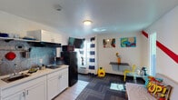 This single-floor studio unit delivers a compact yet fully functional layout with a bold, pop-art-inspired aesthetic. The open-plan main area seamlessly combines the kitchenette, living, and dining zones into one cohesive space. The kitchenette is well-appointed with white shaker-style cabinetry, a black refrigerator, a microwave, a coffee maker, a cooktop, and a sink — all set against a textured metallic backsplash. A wall-mounted flat-screen TV is positioned for easy viewing from the living area. The living space features a daybed-style sofa adorned with comic-themed accent pillows, patterned carpet tiles, and a wall accented with a bold red diagonal stripe. Vibrant pop-art prints and a compact wall-mounted dining table with a glass top and yellow chair complete the look. Natural light filters in through a window dressed with navy blue and white striped curtains, complemented by an under-window HVAC unit. The bathroom is clean and efficiently designed, featuring a walk-in shower with a glass panel, a white vanity with storage, a mirrored medicine cabinet, and tile flooring. The overall ambiance is energetic, creative, and designed to maximize utility within a compact footprint.