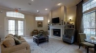 This single-story living space is designed with a traditional aesthetic, centered around a stately white-mantled fireplace that serves as the room's focal point. A flat-screen television is mounted above the mantle, flanked by wall sconces that add warm accent lighting. The fireplace surround features built-in shelving and cabinetry on either side, offering both storage and display space for books and decorative items.

The room benefits from natural light through a glass-paneled entry door and a transom-style window positioned high on the wall, complementing the recessed ceiling lighting throughout. Crown molding adds a refined architectural detail along the upper perimeter of the room.

The flooring is a light-toned tile that transitions seamlessly beneath a large, intricately patterned area rug, anchoring the seating arrangement. Furnishings include a plush sofa, a pair of upholstered accent chairs with nailhead trim, and a tufted wingback chair positioned near the fireplace. A dark wood coffee table with X-frame detailing sits at the center of the arrangement, contributing to the room's classic, well-appointed character.