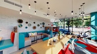 Located on N Interstate Avenue in Portland, Oregon, this single-floor communal space offers a lively, retro-modern aesthetic defined by a bold color palette of teal, red, and gray. The layout is thoughtfully segmented into distinct functional zones while maintaining an open, connected feel throughout.

The lounge area anchors the space with upholstered teal booth seating arranged alongside wood-topped tables, offering semi-private nooks ideal for conversation or focused work. A white-painted brick accent wall serves as a decorative backdrop, adorned with an eclectic arrangement of decorative plates and wall art that adds character and visual interest.

A dedicated workstation nook features a built-in desk with a computer, tucked beside textured herringbone wallcovering and lush hanging greenery, providing a practical workspace within the communal setting. Nearby, an Amazon Hub package locker station offers a convenient, secure mail solution, framed by sculptural white wall décor.

The most striking feature is the floor-to-ceiling curtain wall of black-framed glass along the street-facing edge, flooding the interior with natural light and providing open views of the tree-lined streetscape. Pendant lighting in varying heights adds warmth overhead, while a bold hexagonal patterned carpet grounds the entire space with geometric flair.