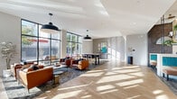 Located at 417 Callowhill Street in Philadelphia's vibrant Callowhill neighborhood, this generously proportioned communal living space offers a thoughtfully designed, single-floor layout that blends modern aesthetics with functional comfort. The space is anchored by a dramatic floor-to-ceiling black-framed glass facade at the entrance, flooding the interior with abundant natural light and offering open views of the surrounding streetscape.

The main living area features warm-toned hardwood flooring throughout, paired with a crisp white ceiling accented by bold pendant lighting. A central communal dining table with mixed seating serves as a focal gathering point, while a curated lounge area with rich cognac leather armchairs and patterned area rugs creates a relaxed, inviting atmosphere.

A distinct lounge nook is defined by slatted wood wall paneling, concrete-look accent columns, and teal upholstered booth seating, offering a more intimate setting within the open plan. Cascading greenery on iron shelving adds organic warmth to this area. An elevator is conveniently integrated into the layout, and a reception counter provides a functional lobby presence. Contemporary artwork and carefully selected furnishings complete the polished, welcoming character of this well-appointed communal space.