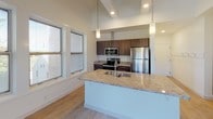 Located on Fulton Street East in Grand Rapids, Michigan, this two-floor residence offers a thoughtfully designed interior that balances contemporary finishes with characterful architectural details. The main level features a well-appointed kitchen with dark espresso cabinetry, stainless steel appliances, granite countertops, and a large peninsula with an undermount sink — all bathed in natural light from generous windows overlooking lush tree canopies. Light wood flooring flows throughout the main living areas, enhancing the bright, airy atmosphere. A half bath on this level features granite countertops, a modern vanity with light wood cabinetry, and clean tile flooring. Ascending to the upper floor, dramatic exposed timber truss beams define the space and add a striking rustic contrast to the otherwise crisp white walls and plush carpeting. A spacious loft-style area with soaring vaulted ceilings and a modern metal railing overlooks the staircase below. Additional rooms on this level feature carpet flooring and multiple windows that invite abundant natural light and outdoor views. A full bath with a dual-sink granite vanity and large format tile flooring serves the upper level. Throughout both floors, the design presents a clean, modern aesthetic with warm natural accents.