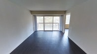 This single-floor space offers a clean, modern layout designed to maximize both comfort and natural light. The main living area is a standout feature, characterized by rich, dark hardwood flooring that contrasts elegantly with crisp white walls and ceilings. A large sliding glass door dominates one wall, flooding the interior with natural light and providing direct access to a private balcony. The balcony overlooks a serene, tree-lined setting, offering a peaceful natural backdrop that enhances the overall ambiance of the space. Sheer curtains frame the expansive glazing, softening the light while maintaining an open, airy feel. The open-plan layout of the living area creates a generous sense of space, with a wide entryway connecting seamlessly to an adjacent room, suggesting versatile use for dining or additional living arrangements. The neutral color palette throughout keeps the interior feeling fresh and adaptable. Structural details such as the clean beam transitions and recessed wall sections add subtle architectural character. Overall, this space strikes a balance between understated elegance and functional design, making it well-suited for a variety of lifestyles.