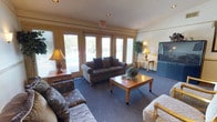 This single-floor lounge area is designed as a relaxed gathering space, offering a well-appointed and functional layout suited for socializing or leisure. The room is anchored by a large bank of windows along one wall, allowing abundant natural light to flood the interior and providing views of the greenery outside. The blue carpeted flooring adds a sense of warmth underfoot and ties the room's neutral color palette together cohesively.

Seating is arranged thoughtfully around a central wooden coffee table, with a cushioned sofa featuring dark throw pillows serving as the focal point. Additional upholstered chairs with decorative leaf-patterned fabric are positioned throughout the room to accommodate multiple occupants comfortably. A large flat-screen television is positioned in one corner, making the space suitable for group viewing. Accent tables with table lamps provide soft supplemental lighting, while a potted decorative tree adds a touch of organic texture to the corners. The textured ceiling and recessed lighting contribute to the room's understated, practical design. An illuminated exit sign indicates this space is part of a larger facility.