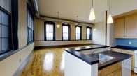This 2-bedroom, 1-bathroom unit showcases a distinctive industrial-loft aesthetic, blending raw architectural details with functional living spaces across a single floor. The open-concept main living area is anchored by warm hardwood flooring that reflects natural light streaming in through a row of large, black-trimmed arched windows. These generously sized windows line the exterior wall, creating a bright and airy atmosphere throughout the day.

Exposed ductwork runs along the ceiling, lending an authentic industrial character to the space, while pendant lights with classic globe and cone-shaped shades hang at varying heights, providing ambient illumination and reinforcing the loft-inspired design.

The kitchen features a wraparound island counter finished in dark laminate, providing ample prep and dining space. A stainless steel sink is integrated into the island, and the white cabinetry base offers clean contrast against the darker countertop. The open layout allows the kitchen to flow seamlessly into the living area, making the space well-suited for entertaining. Overall, the unit combines period-influenced architectural charm with practical, modern conveniences.