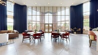 Located at 7475 Flying Cloud Dr in Eden Prairie, MN, this expansive single-floor community lounge offers a generous, open-plan layout designed for both relaxation and social gatherings. The space is immediately distinguished by its dramatically soaring ceilings and a sweeping wall of floor-to-ceiling windows, which flood the interior with abundant natural light and provide unobstructed views of the outdoor courtyard and surrounding greenery.

The main lounge area features a sophisticated seating arrangement centered around a marble-surround fireplace, framed by warm wood slat detailing and complemented by plush leather sofas, accent chairs, and curated artwork. Elegant pendant lighting and a statement chandelier add a refined ambiance throughout.

A dedicated dining area showcases multiple round tables paired with rich red upholstered chairs, set against dramatic floor-length navy drapes that accentuate the vertical scale of the room. Large double wooden entry doors with slatted wood trim provide a grand entrance.

The integrated kitchenette is outfitted with slate-blue cabinetry, a stainless steel refrigerator, a built-in coffee station, and a white quartz island with stylish rattan bar stools — ideal for casual gatherings. Large-format porcelain tile flooring ties the entire space together with a clean, modern aesthetic.