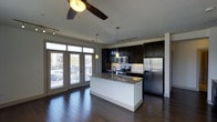 This single-floor space showcases a thoughtfully designed open-concept layout that seamlessly integrates the kitchen and living areas. The kitchen is a standout feature, equipped with stainless steel appliances including a French door refrigerator, electric range, and built-in microwave. Dark espresso-toned cabinetry reaches toward the ceiling, providing generous storage, while a central island with a granite countertop and undermount sink serves as a functional and visual focal point. Pendant lighting hangs above the island, complementing the track lighting and ceiling fan overhead.

Rich, dark hardwood flooring extends throughout the space, adding warmth and continuity. Large French doors with transom windows along one wall invite an abundance of natural light and offer views of the exterior, enhancing the open, airy feel of the interior. The ceiling fan provides both aesthetic appeal and practical climate control. The overall design balances contemporary finishes with functional elements, creating a cohesive and inviting living environment suited for everyday use and entertaining alike.