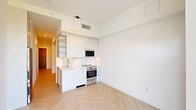 This thoughtfully designed single-floor unit offers a clean, contemporary living environment that maximizes every square foot. Throughout the space, light-toned herringbone-patterned hardwood floors create a visually expansive feel, complemented by crisp white walls and abundant natural light.

The kitchen is a standout feature, showcasing white flat-panel cabinetry extending to the ceiling for ample storage, a marble-look tile backsplash, and white quartz countertops. Stainless steel appliances — including a full-size range, built-in microwave, and refrigerator — provide a full suite of modern cooking amenities. Open shelving adds both functionality and visual interest.

The living area is an open, versatile space with clean white walls and recessed linear LED lighting that casts a warm, even glow. Multiple electrical outlets are thoughtfully distributed throughout, offering flexible furniture and layout options.

The bathroom is finished to a high standard, featuring large-format floor tiles, a soaking tub with a tiled surround, a sleek floating vanity with an integrated sink, a large mirror with LED strip lighting, and polished chrome fixtures throughout.

The hallway connects all areas efficiently, featuring the same herringbone flooring and a distinctive blue-stained wood entry door that adds a bold design accent. Overall, the space presents a cohesive, modern aesthetic with quality finishes at every turn.