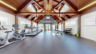 Located at 380 Mather St in Hamden, Connecticut, this fitness center is housed in a single-floor space that makes a striking architectural impression. The room is defined by its dramatic vaulted ceiling adorned with rich, exposed wooden beams and trusses that lend a warm, lodge-like character to the space. Clerestory windows positioned near the roofline, along with a series of larger windows along the walls, flood the interior with generous natural light and provide views of the surrounding community.

The exercise room is comprehensively outfitted to support a wide variety of workout routines. The cardio section features multiple treadmills equipped with individual monitor screens, an upright stationary bike, and an elliptical machine. For strength training, the space includes a cable/functional trainer machine, adjustable weight benches, a full rack of dumbbells, barbells with weight plates, kettlebells, and a medicine ball storage rack. A punching bag and wall-mounted resistance equipment round out the functional training options. Storage cubbies and organizational fixtures keep the space tidy and accessible.

The flooring throughout is durable rubber gym matting in a dark tone, providing cushioning and safety underfoot. The walls are painted in a calming blue on one side and crisp white on the other, complementing the warm wood overhead and creating a clean, motivating environment for exercise.