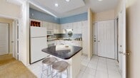 This single-floor residence offers a thoughtfully designed open-concept layout that seamlessly connects the kitchen, dining, and living areas. The kitchen is equipped with white cabinetry, a full suite of white appliances, and a central island featuring a dark countertop and bar seating — ideal for casual dining or entertaining. Recessed lighting and tile flooring enhance the kitchen's clean, functional aesthetic, while a soft blue accent wall adds a touch of color.

The open living and dining area flows naturally from the kitchen, featuring carpeted flooring, a ceiling fan, and sliding glass doors that lead to an outdoor space, filling the room with natural light. A second living or lounge area provides additional flexible space, complete with recessed lighting and room for a home office setup.

The primary bedroom is generously sized with plush carpeting, recessed lighting, and a window dressed with bold striped curtains. The en-suite bathroom includes a wide vanity with a large mirror, ample cabinetry, and a soaking tub, all finished in crisp white tile. A second bathroom features a glass-enclosed walk-in shower with mosaic tile flooring. The second bedroom mirrors the primary in comfort, offering soft carpet and recessed lighting throughout.