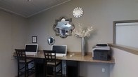 This single-floor space features a well-appointed workstation area designed to balance functionality with aesthetic appeal. The built-in granite-topped desk spans the width of the wall, providing ample workspace and accommodating multiple computer stations, complete with desktop monitors and a printer for practical use. Dark wood chairs complement the warm tones of the granite surface, creating a cohesive and grounded look. The walls are finished in a soft neutral gray, lending the space a calm and professional atmosphere. Decorative elements elevate the design, including an ornate mirrored wall piece with a floral geometric shape, a sunburst mirror, and a smaller diamond-shaped mirror, all arranged artfully on the wall. A tall silver vase holding elegant white branches adds a refined, contemporary touch. The ceiling features a light wood panel finish, adding warmth and texture overhead. Overall, the space strikes a balance between a productive work environment and tasteful interior design, making it both inviting and practical for everyday use.