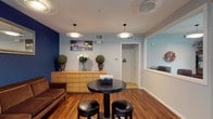 This leasing office is thoughtfully designed to create a professional and inviting atmosphere for prospective residents. The single-floor layout is unified by rich, warm hardwood flooring throughout, providing a polished foundation that complements the space's contemporary aesthetic.

The main reception area features bold navy blue accent walls contrasted against light gray surrounding walls, creating a visually dynamic environment. Pendant lighting fixtures with circular shades provide consistent, well-distributed illumination throughout. A comfortable brown sofa offers seating for visitors, accompanied by a round dark bistro table with black leather stools for more casual interactions. A light wood dresser along one wall serves as a display surface, decorated with topiary plants and floral arrangements, adding a welcoming touch.

A dedicated beverage station, complete with a water cooler and coffee maker, is conveniently positioned for guest use. A wall-mounted mirror and framed artwork add decorative character to the space.

A glass-partitioned sliding door separates the main area from a private meeting room, which features a formal table with upholstered chairs and direct access to an outdoor area through sliding glass doors. A restroom is accessible from the main corridor, and natural light filters in through window blinds, enhancing the overall ambiance.