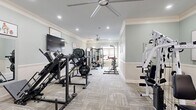 Located at 445 Warfield Blvd in Clarksville, Tennessee, this fitness center offers a generously sized, single-floor layout designed to accommodate a comprehensive workout experience. The space is finished with refined architectural details, including crown molding, chair rail wainscoting, and soft sage-green walls that create a clean, welcoming ambiance throughout.

The flooring consists of durable patterned carpet tiles, providing comfort underfoot during workouts. Recessed lighting and multiple large ceiling fans ensure the space remains well-lit and well-ventilated. Natural light flows in through windows at the far end of the room, where glass doors provide an exit to the outdoors.

The fitness floor is thoughtfully arranged to maximize usability. One end of the room houses cardio equipment, including multiple treadmills and a stair climber. Moving through the space, users will find an array of strength training stations, including a multi-function cable machine, leg press, weight benches with barbell racks, and a cable crossover system. Additional accessories such as resistance bands, medicine balls, and yoga mats are neatly organized near the entrance. Wall-mounted flat-screen televisions and mirrors are positioned throughout to enhance the workout experience. The layout allows for comfortable movement between stations, making this fitness center a functional and attractive amenity.