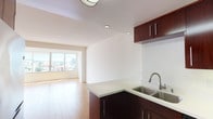This upper-floor residence offers a bright, open layout with clean lines and a neutral palette throughout. The spacious living area is finished with warm hardwood flooring and is bathed in natural light from large floor-to-ceiling windows that frame panoramic city and hillside views. A sliding glass door leads to a private balcony, extending the living space outdoors.

The kitchen is thoughtfully designed with rich dark wood cabinetry, light-colored countertops, a double stainless steel sink, and a full suite of stainless steel appliances including a refrigerator, range, microwave, and dishwasher. A peninsula counter separates the kitchen from the living area, creating a seamless and functional open-plan layout.

The bedrooms are generously sized, carpeted in a neutral grey tone, and feature built-in closets with ample storage. The layout provides a sense of privacy, with the bedrooms positioned away from the main living areas. An additional room with carpeting and closet access offers flexibility for various uses.

The bathroom includes a full soaking tub with clean white tile surround, maintaining the residence's consistent, understated aesthetic. Recessed lighting throughout adds a polished finish to each room, while white walls create a bright, airy ambiance across the entire space.