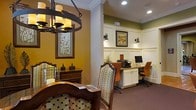 This single-floor space combines classic elegance with practical functionality, creating an inviting environment for residents and guests alike. The dining area exudes warmth with its rich hardwood furniture, including ornately carved chairs upholstered in patterned fabric, and a striking wrought-iron chandelier fitted with candle-style lights that casts a warm, ambient glow. Earthy golden-yellow walls, accented by crisp white crown molding, lend a refined yet cozy character to the space. A traditional wooden sideboard with decorative accents adds to the classic aesthetic. Adjacent to the dining area, a dedicated business center provides a productive workspace, thoughtfully designed with a built-in white cabinetry workstation, recessed and wall-mounted task lighting, and computers available for use. The space features plush carpeting in a deep burgundy tone, adding warmth underfoot, while orange ergonomic chairs offer comfortable seating. Olive-green upper walls trimmed with white wainscoting paneling lend the business center a distinguished, professional feel. Framed botanical artwork displayed throughout ties the two areas together cohesively, resulting in a space that is both functional and aesthetically refined.