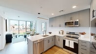 Located at 801 N Orange St in Wilmington, Delaware, this thoughtfully designed one-bedroom, one-bathroom residence offers a sleek, modern aesthetic across a single floor. The open-concept living and kitchen area serves as the heart of the home, bathed in natural light from dramatic floor-to-ceiling windows that frame views of the surrounding neighborhood and a community pool below. The living space features light hardwood flooring and a clean, neutral palette that enhances its bright and airy atmosphere. The kitchen is well-appointed with gray flat-front cabinetry extending to the ceiling, stainless steel appliances including a refrigerator, electric range, and over-the-range microwave, and a white quartz waterfall-edge island with bar seating and a pendant light above — ideal for both cooking and entertaining. The bedroom is a serene retreat, benefiting from its own floor-to-ceiling corner window that floods the room with light and offers outdoor views. Light hardwood flooring continues throughout. The bathroom features large-format slate tile flooring, a walk-in shower with marble-style tile surround, a wood-toned vanity, and a frameless mirror. A dedicated closet completes this efficient and stylish residence.