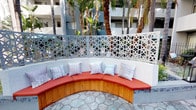 This inviting outdoor courtyard is thoughtfully designed to blend comfort and contemporary style within a lush, open-air setting. The centerpiece of the space is a gracefully curved, built-in bench upholstered in bold coral-red cushions and accented with an assortment of silver and pastel throw pillows, providing generous seating for relaxing or socializing. The bench is framed by a striking decorative screen featuring an intricate geometric lattice pattern in crisp white, which serves as both a visual boundary and a design statement, allowing filtered views of the surrounding palm trees and multi-story building façade beyond.

The flooring throughout the courtyard consists of large, irregularly shaped stone pavers in soft blue-grey tones, adding a natural, organic texture underfoot. Towering palm trees are positioned throughout the space, providing shade and reinforcing the resort-like atmosphere. Additional seating areas are arranged further into the courtyard, including lightweight chairs and small bistro-style tables shaded by a teal market umbrella, ideal for casual dining or conversation.

Lantern-style light fixtures are placed along the pathways, enhancing the ambiance during evening hours. Lush plantings and shrubs are interspersed throughout, softening the hardscape and adding layers of greenery. The overall ambiance is that of a well-appointed, design-forward outdoor lounge space with a warm, welcoming character.