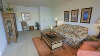 This single-floor space showcases a relaxed, tropical aesthetic throughout its open living area. The main living room is carpeted and centered around a floral-patterned sofa paired with a decorative dark-wood and wicker coffee table, anchored by a warm terracotta area rug. A rattan accent chair and matching side table add to the coastal, laid-back character of the room. Two framed botanical prints depicting palm trees adorn the wall above the sofa, reinforcing the tropical theme. A table lamp provides warm ambient lighting, complementing the natural light that filters in through a window on the adjacent wall. A potted tropical plant adds a touch of greenery, enhancing the breezy atmosphere. The entryway is clearly defined by a white door, creating a natural transition into the living space. The neutral-toned walls and light-colored carpet keep the space feeling open and airy. Overall, the space offers a cohesive, resort-inspired ambiance that feels both inviting and casually comfortable, suited for relaxed everyday living.