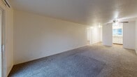 Located at 10728 NE Halsey St in Portland, Oregon, this single-floor, three-bedroom, one-bathroom residence offers a practical and comfortable layout. The living area is generously proportioned, featuring plush carpeting and a textured ceiling, with a sliding glass door that opens to a private balcony, allowing natural light to filter in and providing views of the surrounding greenery. The kitchen is positioned adjacent to the living space and features white cabinetry, a refrigerator, and a countertop workspace, offering a functional and efficient cooking area. The primary bedroom is notably spacious, providing ample room for furnishings and day-to-day comfort. All three bedrooms are carpeted and include closets, with two of the rooms featuring sliding closet doors for convenient storage. The secondary bedrooms benefit from windows that invite natural light and views of outdoor greenery. A connecting hallway links the bedrooms to the shared bathroom, which is finished with warm wood-look flooring, a combined shower and bathtub, and clean white fixtures. The overall design is clean and neutral throughout, with light-toned walls creating a bright and versatile living environment suitable for a range of residents.